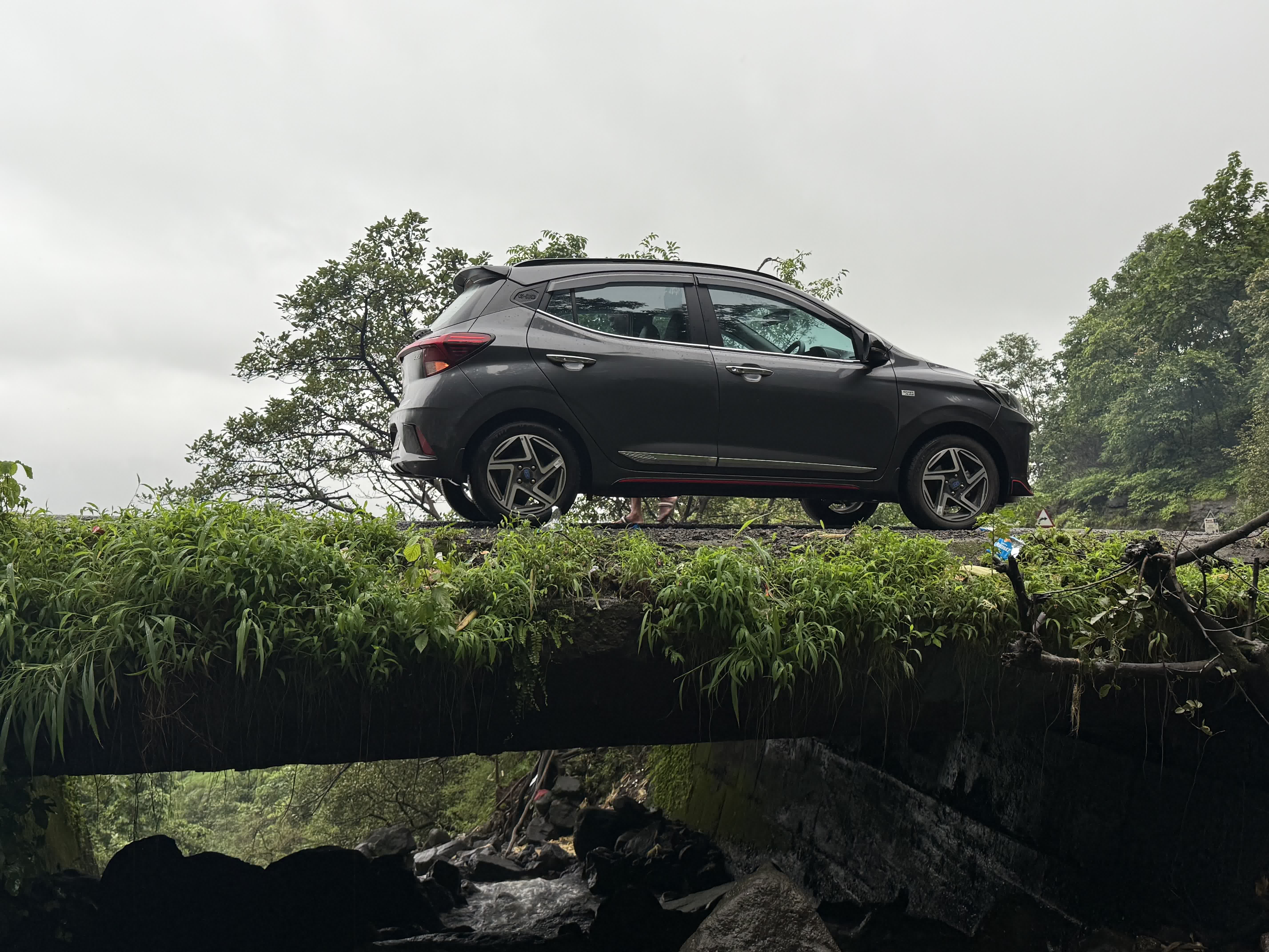 The car parked side-on on a mossy stone bridge over a monsoon stream