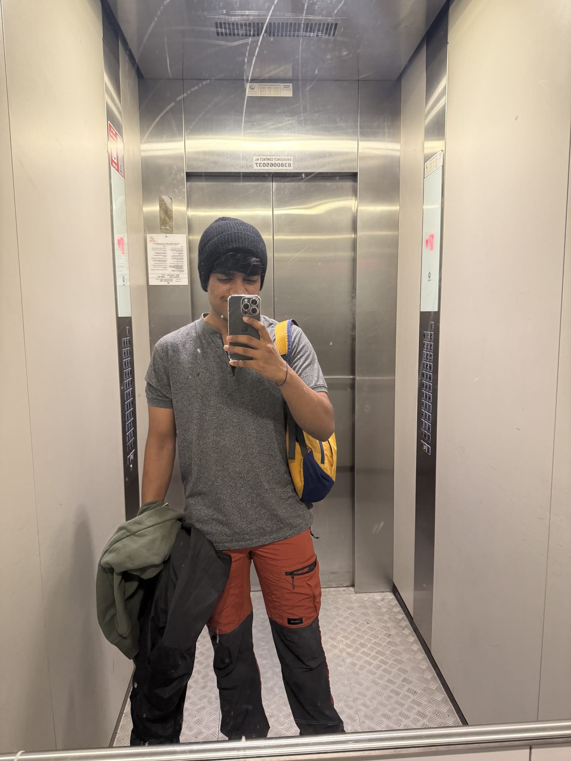 Mirror selfie in a lift, geared up with a beanie and backpack before the trek